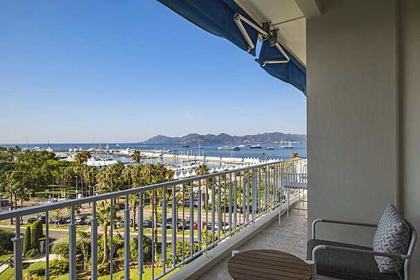 Balcony view overlooking a sunny harbor filled with boats and palm trees, with mountains in the distance under a clear blue sky.