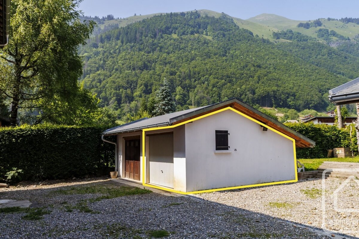 A white garage with two doors and a window, outlined in yellow, sits on a gravel drive with green mountains and trees in the background.