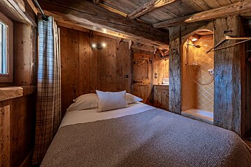 A cozy, rustic wooden bedroom features a bed, window with plaid curtain, and an integrated shower with warm yellow patterned tiles.