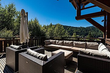 An inviting outdoor terrace with comfortable seating, offering panoramic views of forested mountains under a bright blue sky.