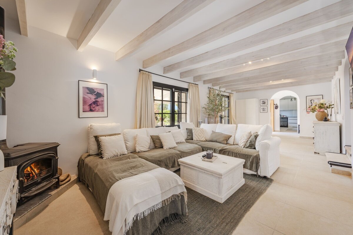 A bright, cozy living room featuring an L-shaped sofa, wood-burning stove, whitewashed beamed ceilings, and an arched doorway, decorated in neutral tones.
