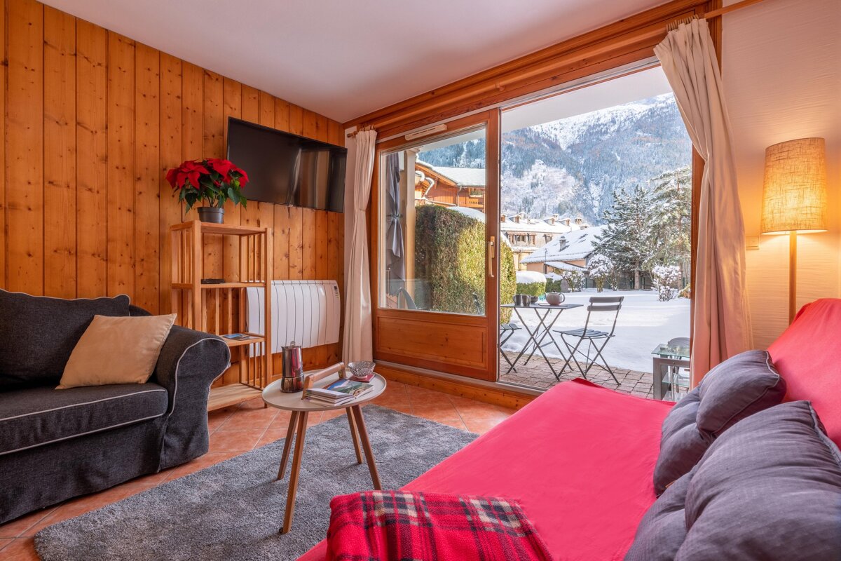 A cozy, wood-paneled living room with sofas and a large sliding door opening to a snow-covered patio and majestic mountains. Perfect winter chalet.