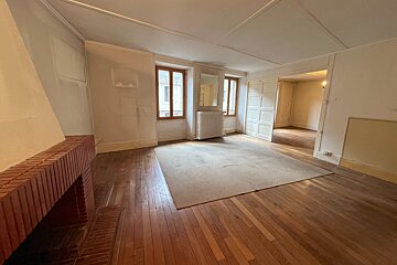 An empty room with wooden floors, a central rug, and a brick fireplace. Windows allow light in, and an open doorway leads to another room.
