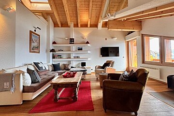 Cozy chalet living room featuring wooden vaulted ceilings, a large sofa, a wooden coffee table on a red rug, and armchairs.