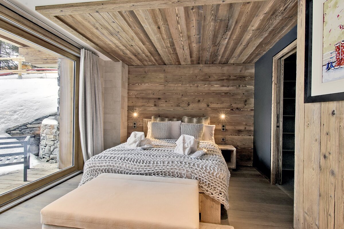 A cozy, wood-paneled bedroom with a comfy bed, warm lighting, and a large window revealing a snowy winter landscape.