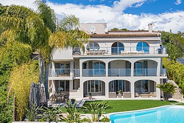A grand, multi-level villa with balconies, a swimming pool, and tropical plants under a blue sky.