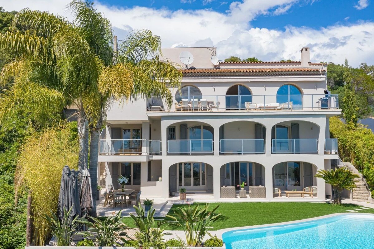 A luxury villa with arched terraces, a swimming pool, and a green lawn under a partly cloudy sky.