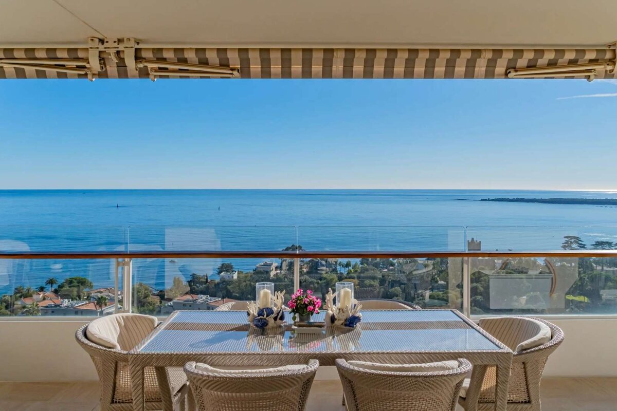 Sunny balcony with an outdoor dining set and a panoramic view of the deep blue sea and coastal landscape below.