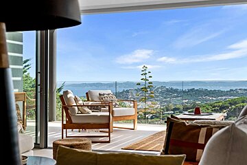 A modern indoor-outdoor space with panoramic sea and coastal views. Stylish chairs on a balcony overlook the beautiful landscape under a blue sky.