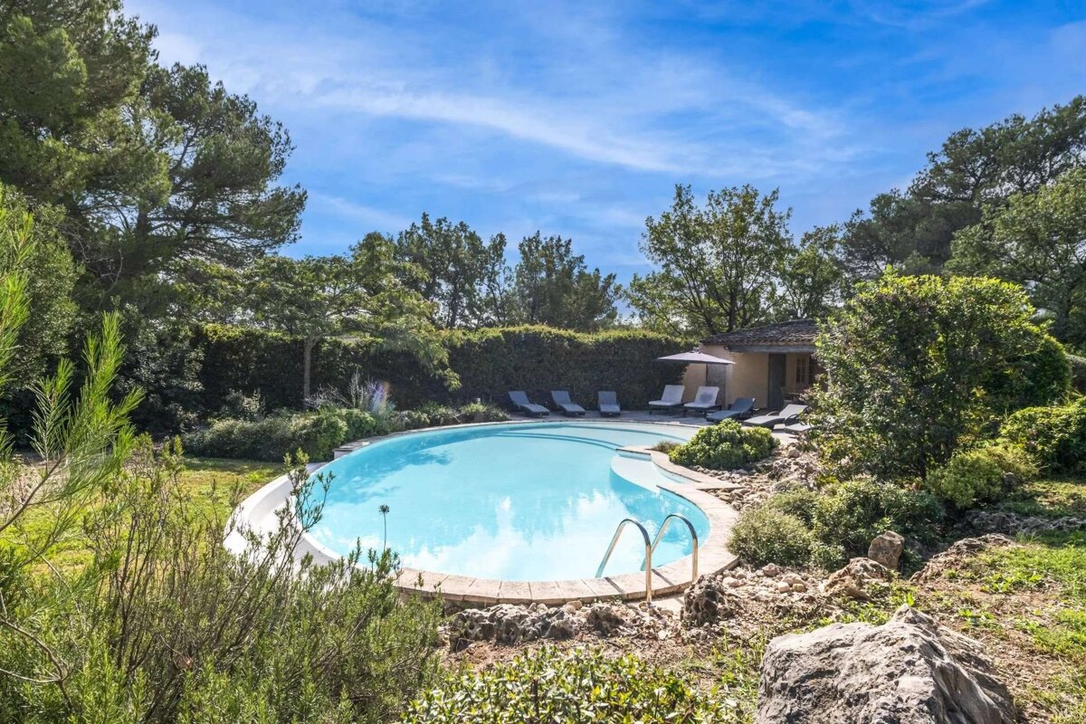 A beautiful curved swimming pool surrounded by lush green trees and bushes under a clear blue sky. Lounge chairs and a small building are nearby.