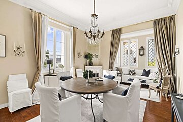An elegant room featuring a round dining table with white slipcovered chairs, a white sofa, and a chandelier, all in warm neutral tones.
