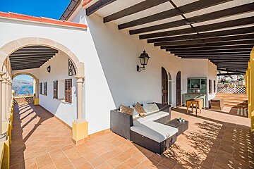 An inviting Mediterranean patio with white walls, terracotta floor, exposed beams, a comfy sofa, and an arched view of a sunny blue bay.