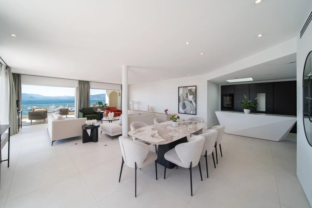 Luxurious open-plan room with modern white decor, dining area, integrated kitchen, and a spacious balcony offering stunning sea views with boats.