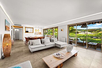 A modern, open-concept living room with a gray sectional, dining area, and kitchen, opening to a balcony with lush green views.