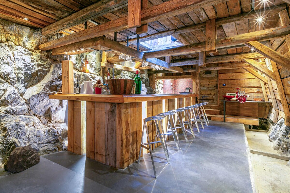 A cozy, rustic bar in a stone and wood cellar with exposed beams, bottles, glasses, and modern stools, all warmly lit.
