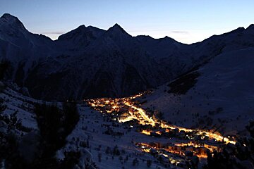 A cozy mountain village glows brightly at twilight, nestled among dark, snow-covered peaks.