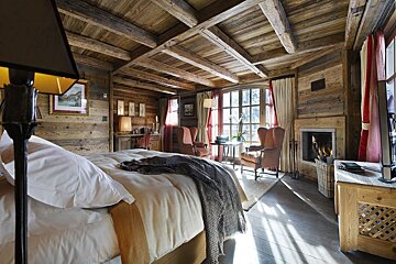 A cozy, rustic wooden chalet bedroom with exposed beams, a large bed, fireplace, and a sunlit sitting area by large windows with red drapes.