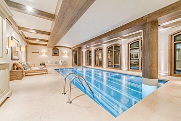 Luxurious indoor swimming pool with warm lighting, wood-beamed ceilings, arched windows, and a comfortable lounge space.