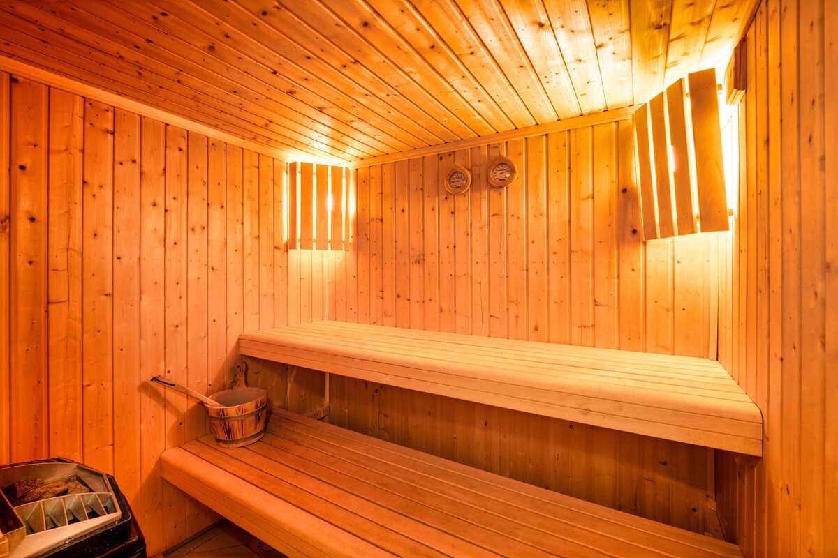 A warm, glowing wooden sauna room with two benches, a bucket, heater, and gauges on the wall, illuminated by soft light.