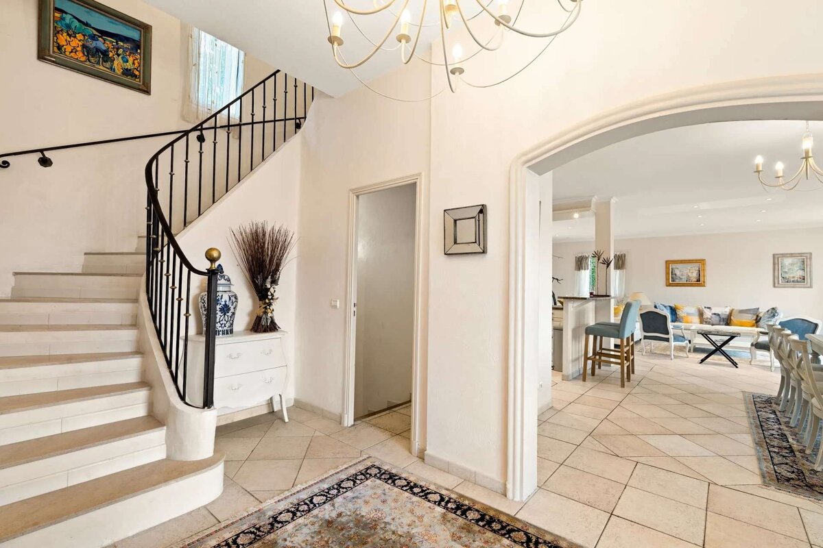 A grand foyer with a curved staircase, elegant chandelier, and decorative rug, leading through an archway to a bright living and dining area.