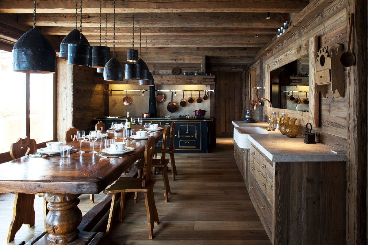 A warm, rustic wooden kitchen and dining room with a long table set for dinner, a traditional stove, hanging copper pots, and farmhouse sinks.