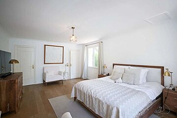 A bright white bedroom with a wooden bed, white chevron duvet, wooden floor, and modern light fixtures. A white armchair and dresser complete the look.