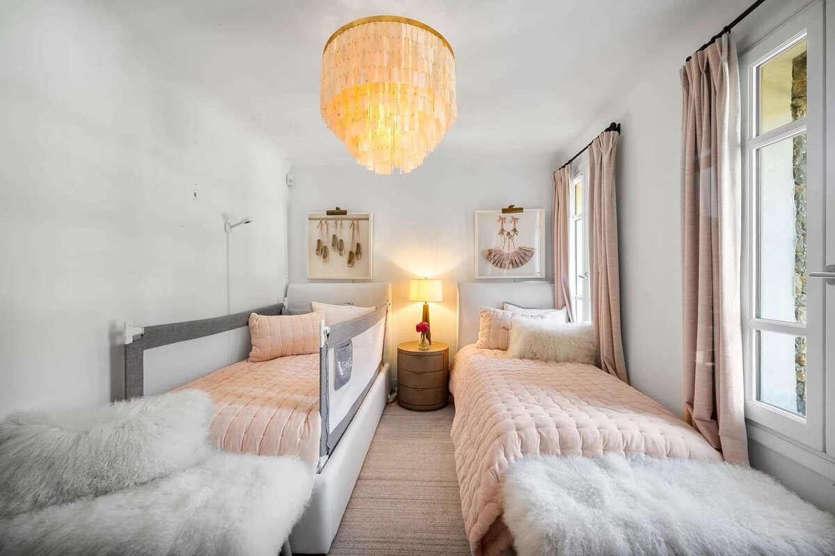 A bright, elegant bedroom with two single beds, soft pink bedding, white fluffy throws, and ballet-themed decor. A statement chandelier lights the space.