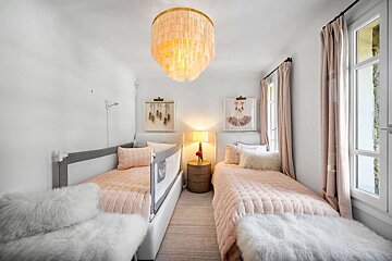 A bright white bedroom features two twin beds with blush pink bedding, fuzzy white throws, and ballet art, illuminated by a large chandelier.