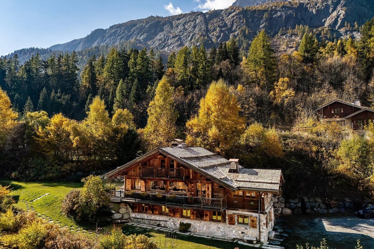 Luxurious wooden chalet with balconies, nestled amidst vibrant autumn trees and towering mountains. Another cabin is visible further up the hill.