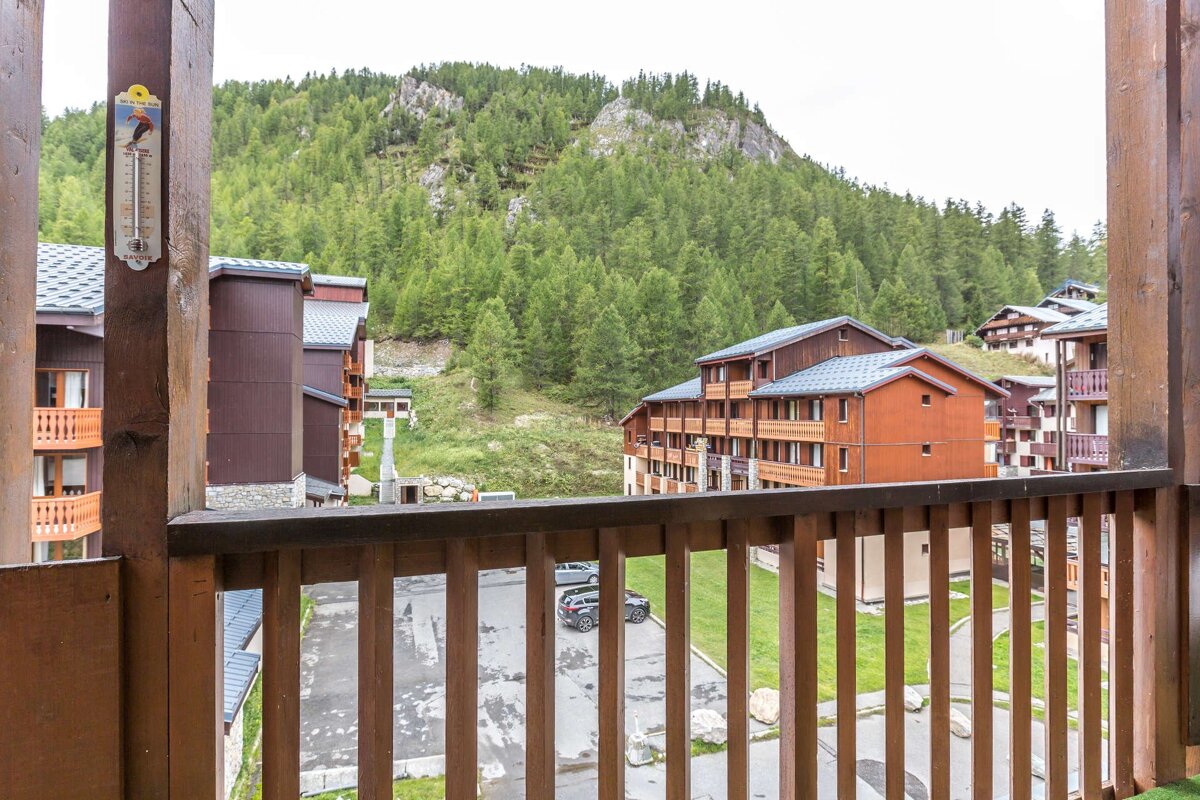 Balcony view of a mountain ski resort with traditional wooden buildings, a forested slope, and a thermometer on a post.