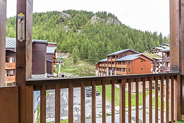 A balcony view of a mountain resort featuring wooden buildings, a dense forest, and a cloudy sky. A skier thermometer hangs on the left railing post.