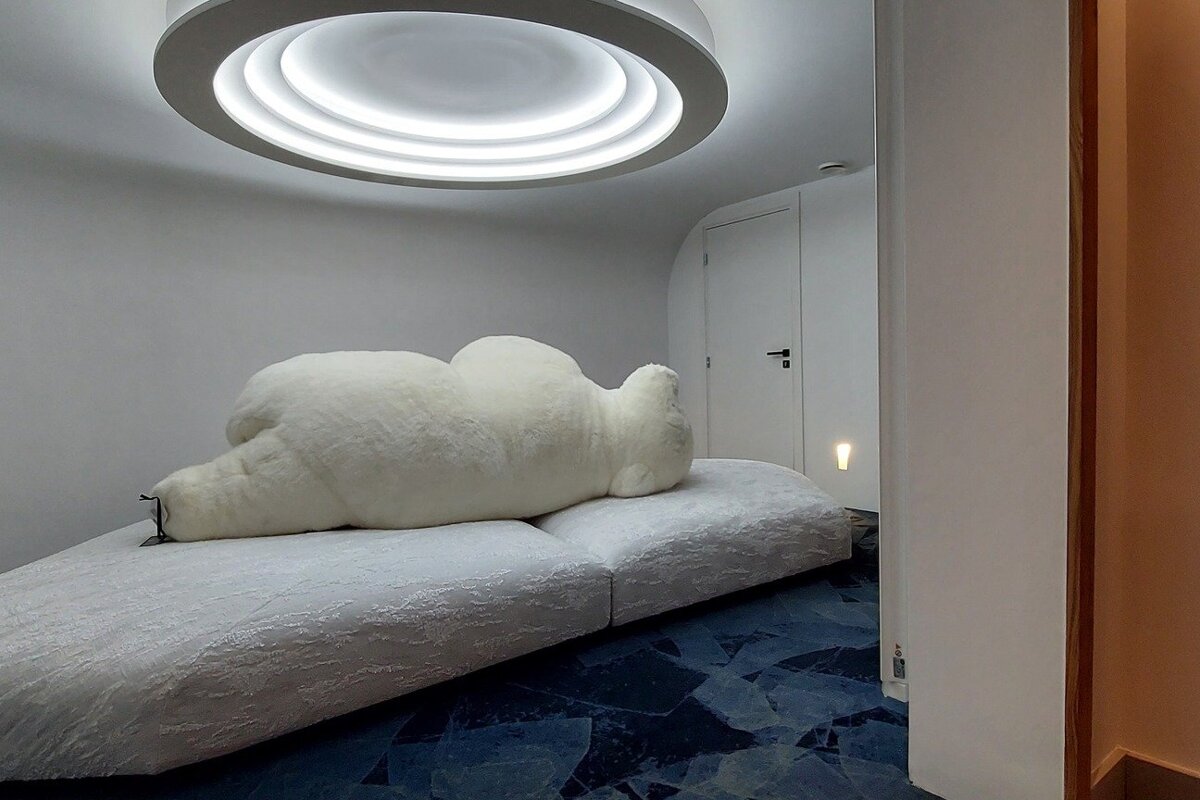A minimalist room features a large, fluffy white bear-like form resting on a low white couch, under a circular ceiling light, with a blue mosaic floor.