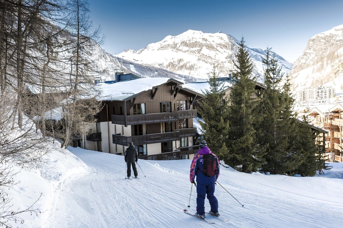 Two skiers glide down a snowy slope at a mountain resort, surrounded by chalets, pine trees, and majestic snow-capped peaks under a blue sky.