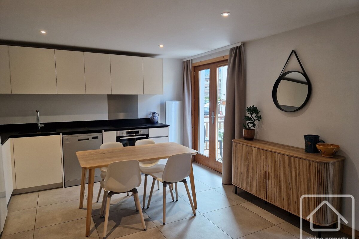 Modern open-plan kitchen and dining area with a wooden table, white chairs, a stylish cabinet, and a round mirror on the wall.