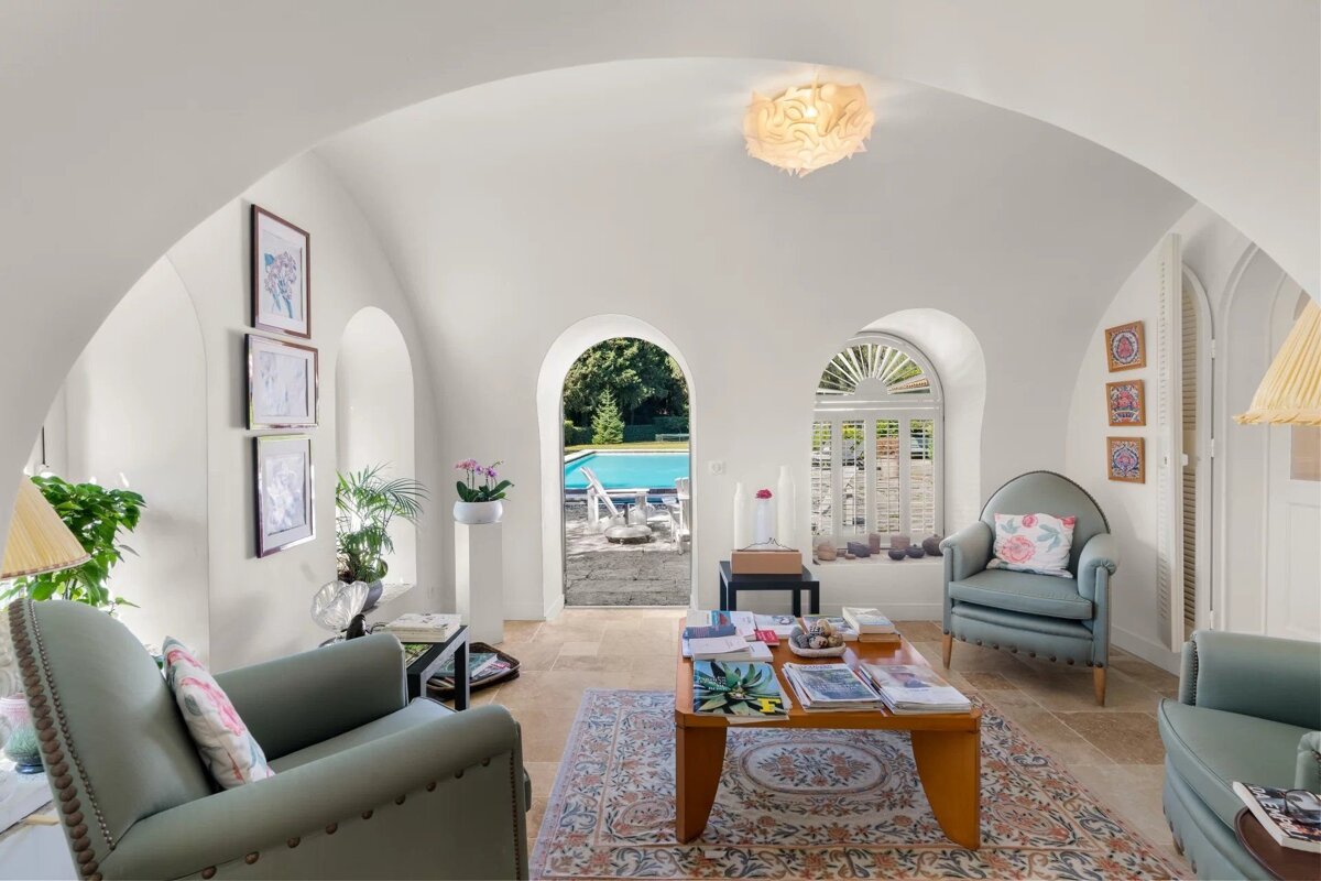 Bright, white living room with arched ceilings, blue armchairs, and an open doorway revealing a sunny patio and swimming pool.