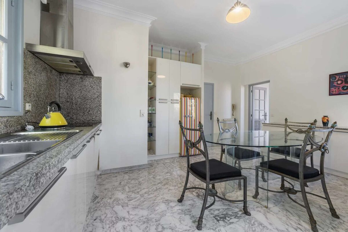 A bright kitchen and dining area features granite countertops, a yellow kettle, white cabinets, and a striped retro fridge, alongside a glass dining table with ornate metal chairs, all set on a marble