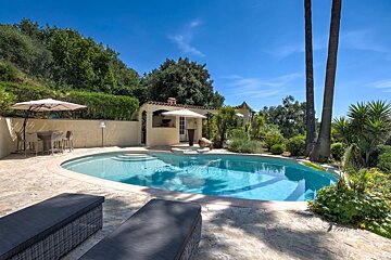 A beautiful backyard patio with a curved swimming pool, lounge chairs, dining area, and pool house surrounded by lush greenery.