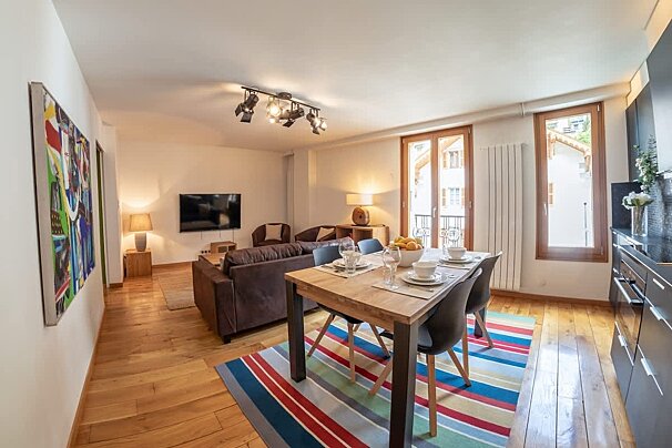 Modern open-plan apartment featuring a wooden dining table on a colorful striped rug, living area with a brown sofa, and warm wooden floors.