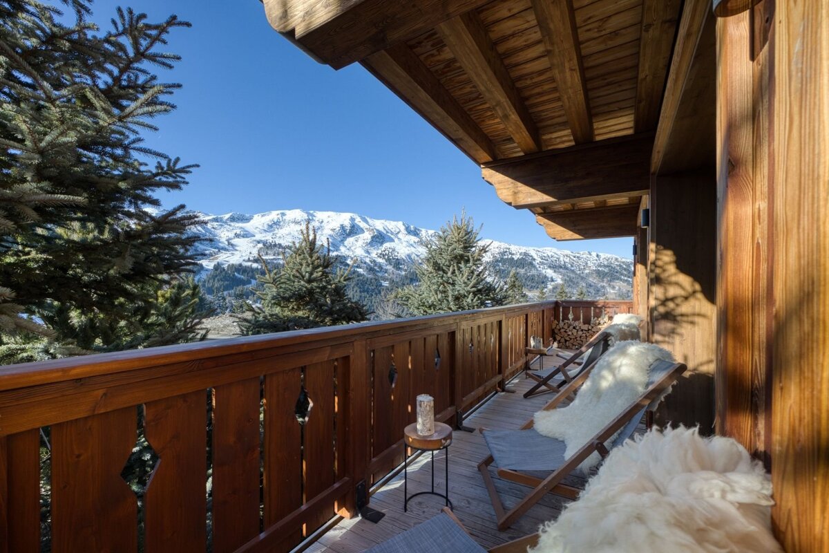 Wooden chalet balcony with cozy fur-draped lounge chairs, overlooking a stunning snowy mountain landscape.