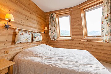 A cozy wooden bedroom with a large bed, two windows offering mountain views, and rustic decor including decorative pillows and wall lamps.