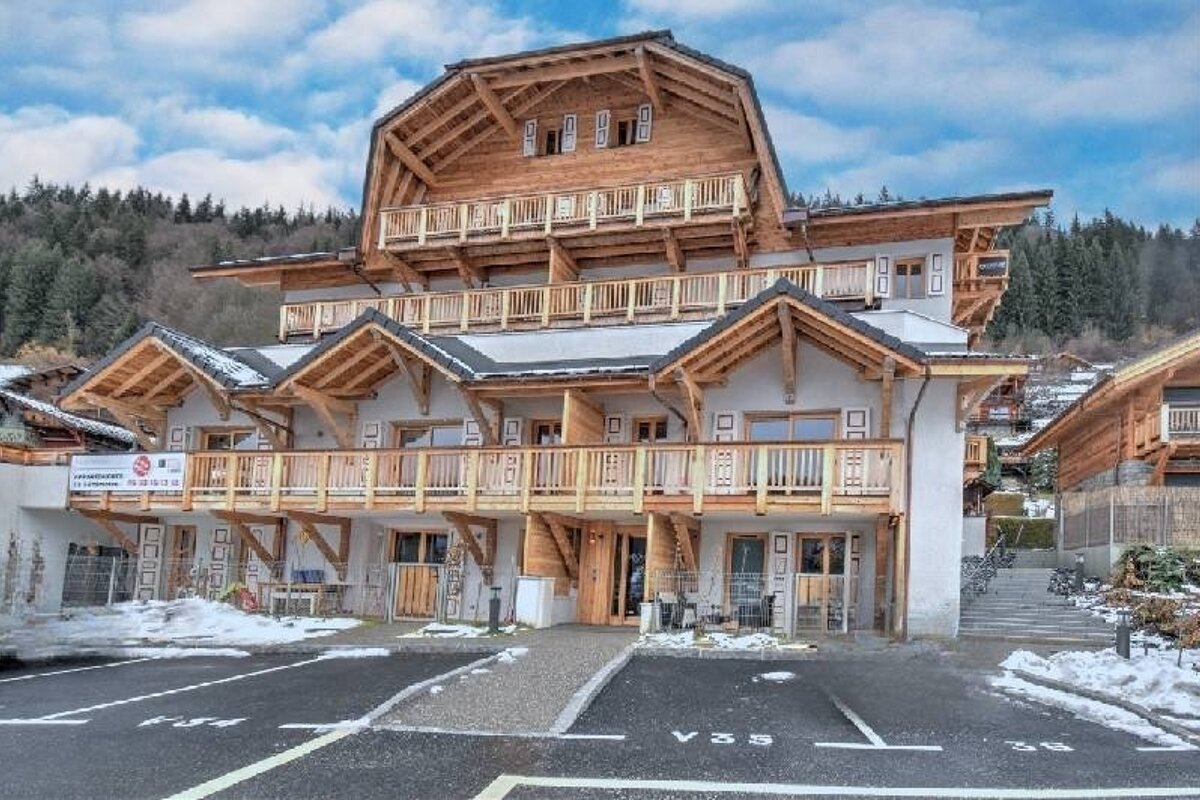 A multi-story wooden alpine chalet building with a parking lot in front, set against a forested hillside under a cloudy sky.
