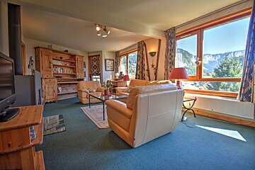 Cozy living room with tan leather armchairs, a wooden cabinet, teal carpet, and a large window looking out at mountains.