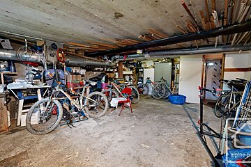 A cluttered basement or storage room with a concrete floor, filled with several bicycles, tools, shelving, and various items. Exposed pipes and wood beams are visible on the ceiling.