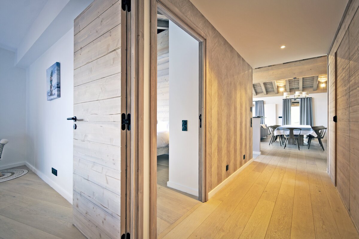 Modern chalet hallway with light wood paneled walls, hardwood floors, and doors leading to a dining room in the background.