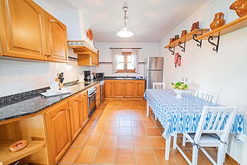 A bright, spacious kitchen with light wood cabinets, dark countertops, terracotta floor tiles, and a dining area featuring a blue patterned tablecloth.