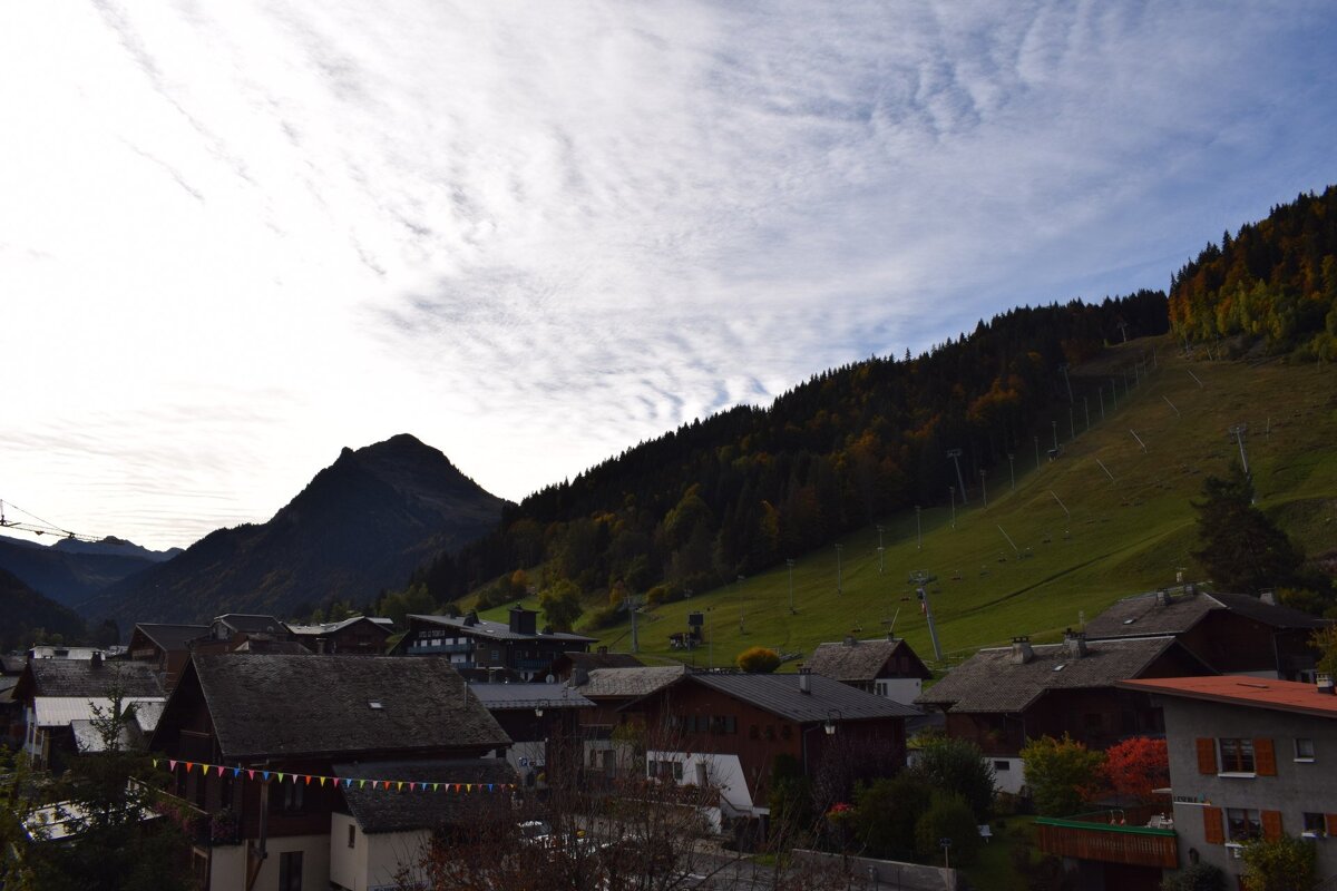 A mountain village with dark-roofed houses lies beneath a steep, forested ski slope and a towering dark peak. A patterned white and blue sky is above.
