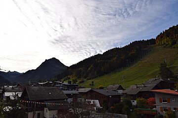 A mountain village of chalets beneath a dark peak, with autumn trees, a green ski slope with lifts, and a cloudy sky overhead.