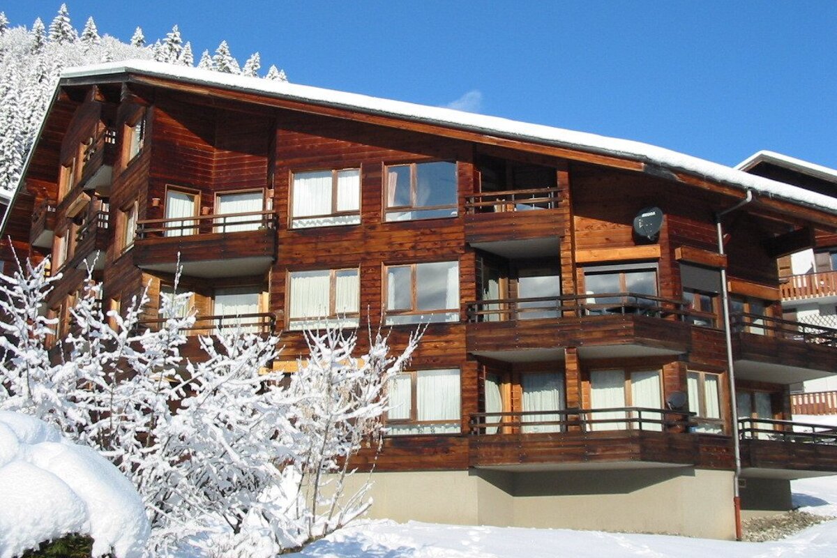 A multi-story wooden chalet with balconies, surrounded by snow and snow-covered trees under a clear blue sky.