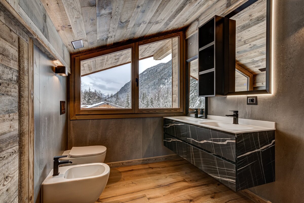 A modern rustic bathroom featuring a black marble vanity, sleek fixtures, and wooden textures, with a large window overlooking snowy mountains.