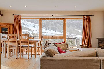 A rustic chalet living room with a wooden dining table and sofa, featuring large windows that offer a picturesque snowy mountain landscape view.